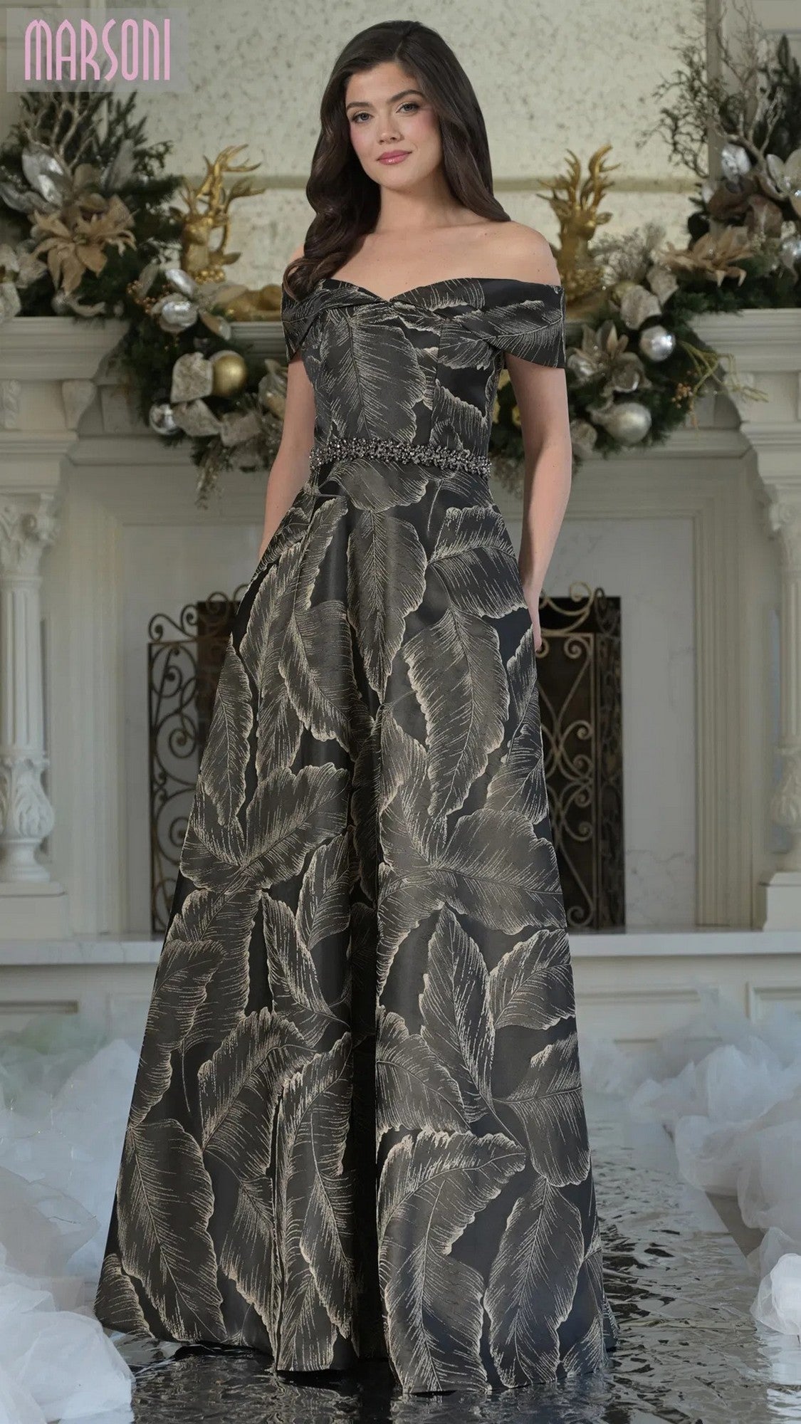 A woman poses indoors in front of a decorated white mantel, wearing the Marsoni MV1265 long formal dress in black—an off-the-shoulder evening gown with a leaf pattern and beaded waistline that exudes timeless elegance.